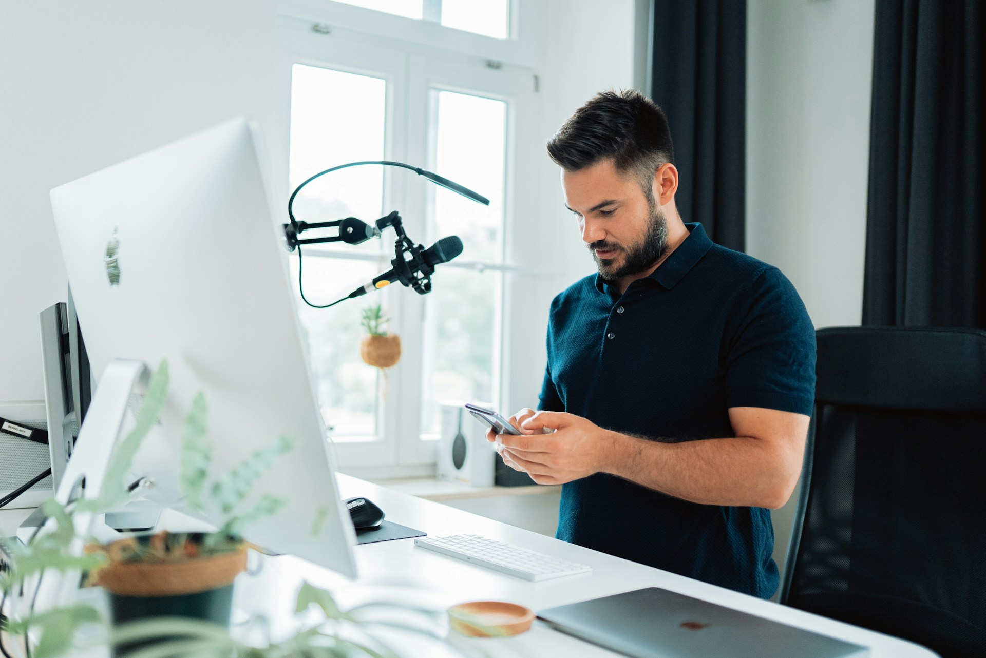 Influencer standing at desk in front of computer and microphone holding a phone. 